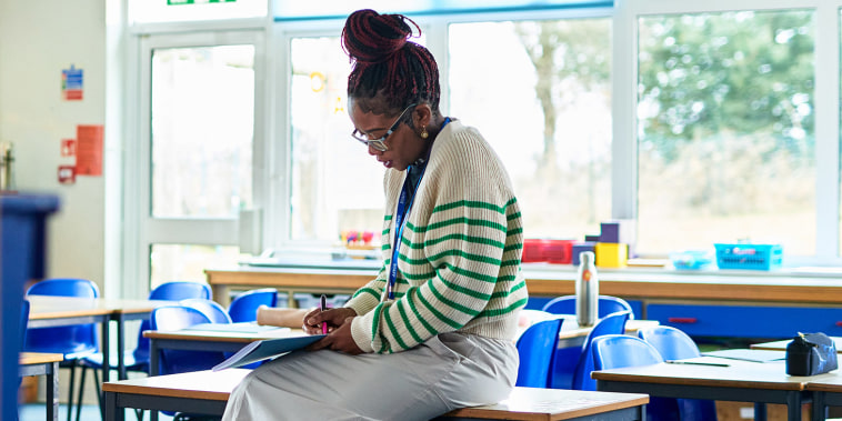 Mid adult female teacher sitting on desk with classroom door open